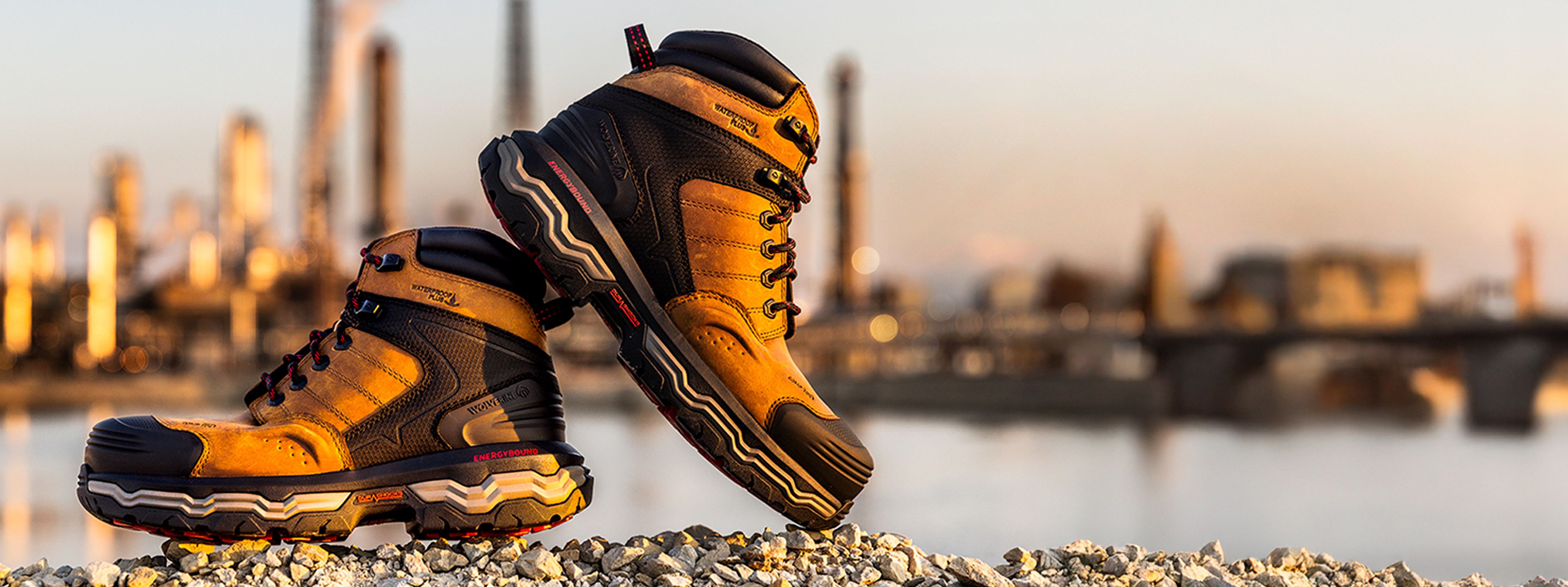 Dark Tan Wolverine Infinity Alpha boot on gravel beside a river with out of focus factories in the background.