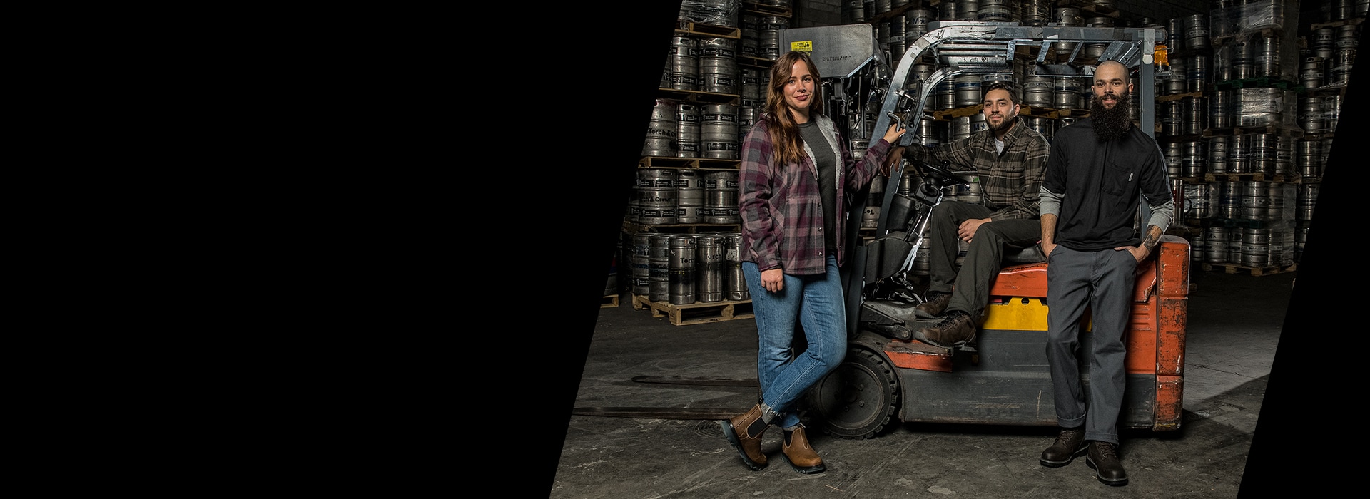 3 friends, wearing Wolvering apparrel and boots, posing by a forklift in a brewery.