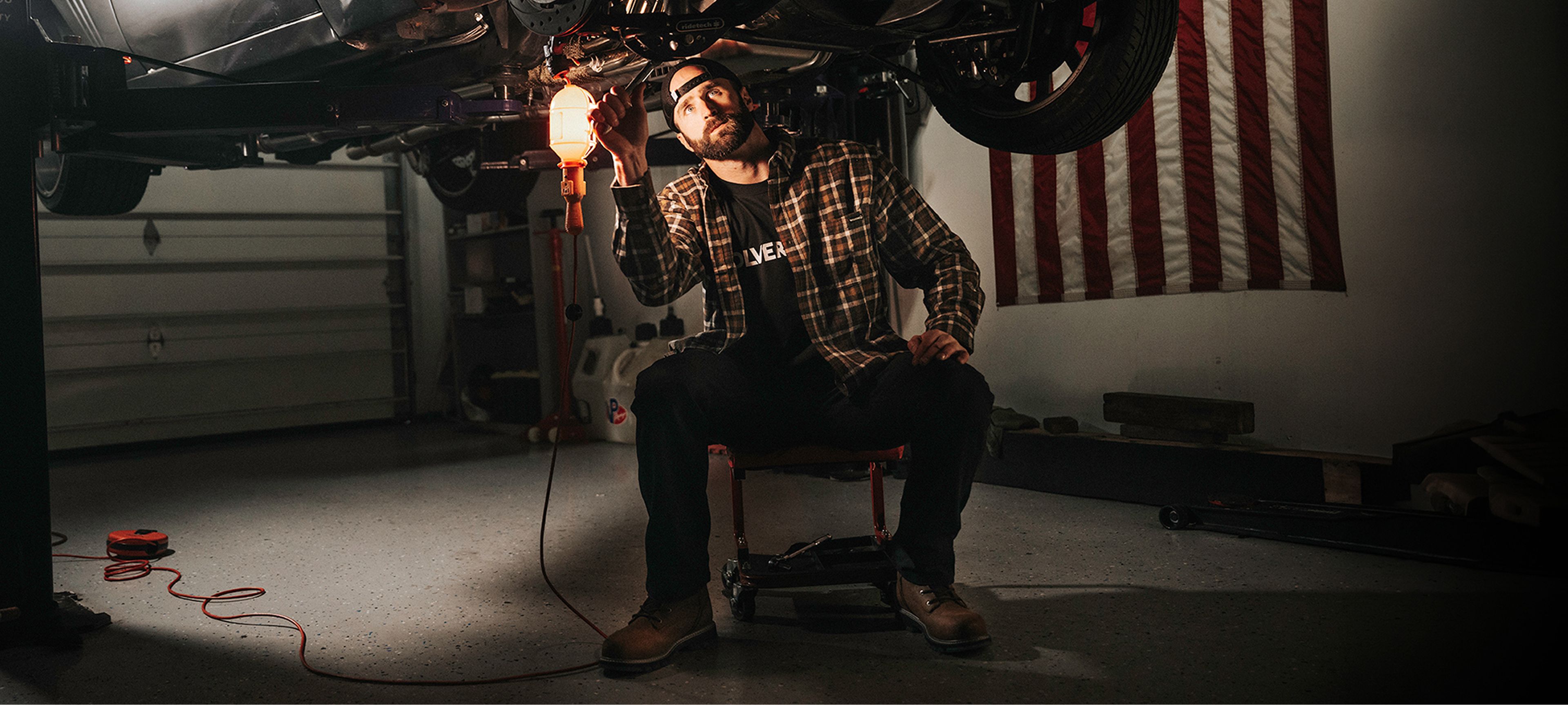 Sitting on a chair, working underneath a car using a flashlight.
